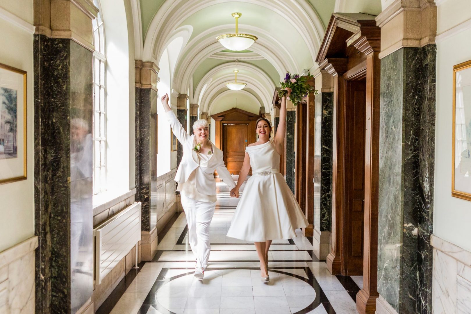 Two brides walking with arms raised at Islington town hall