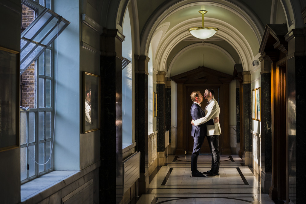 A wide angle shot of two grooms embracing on their wedding day in a corridor 
