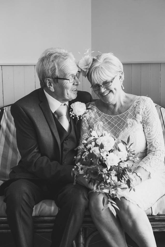 A bride and groom in their 70s laugh as they sit together on a chair