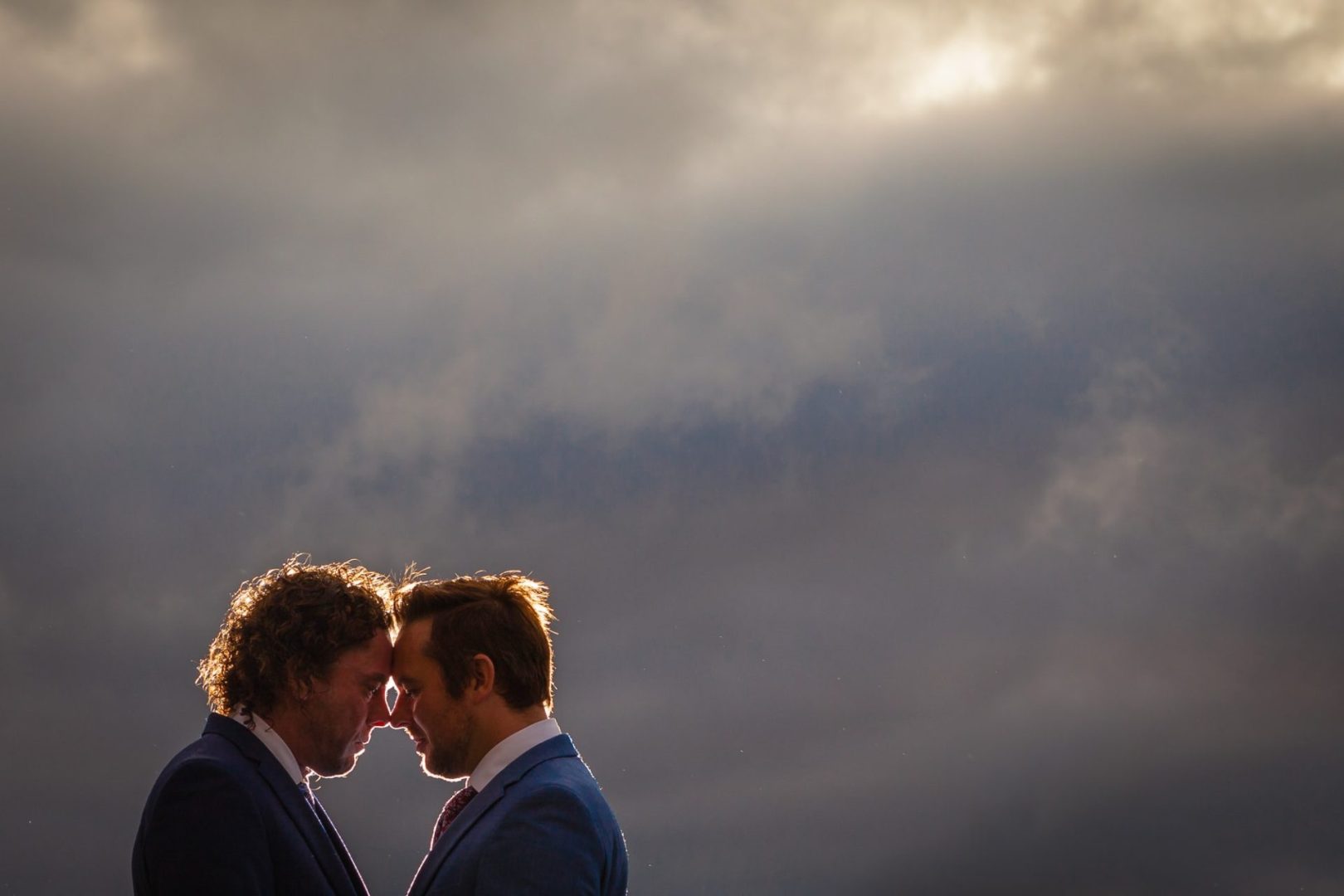 Two grooms heads together under a cloudy sky