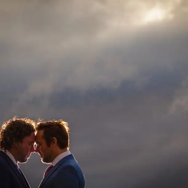 The head and shoulders of two grooms with foreheads together under a cloudy sky