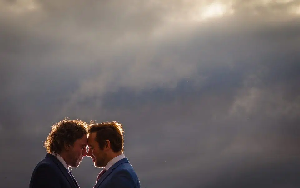 The head and shoulders of two grooms with foreheads together under a cloudy sky