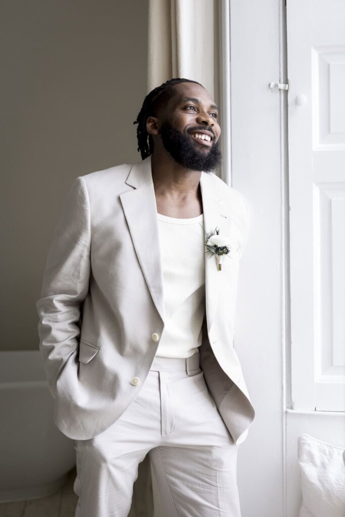a groom smiling whilst looking out of the bedroom window at butley priory