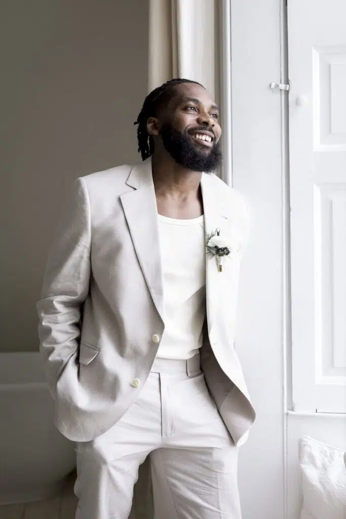 a groom smiling whilst looking out of the bedroom window at butley priory