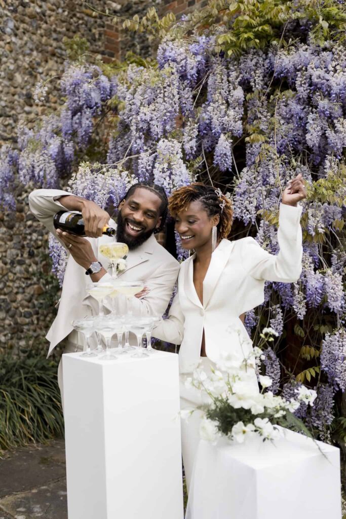 a bride and groom cheer as they pour champagne into a champagne tower in front of the wisteria at Butley Priory Essex