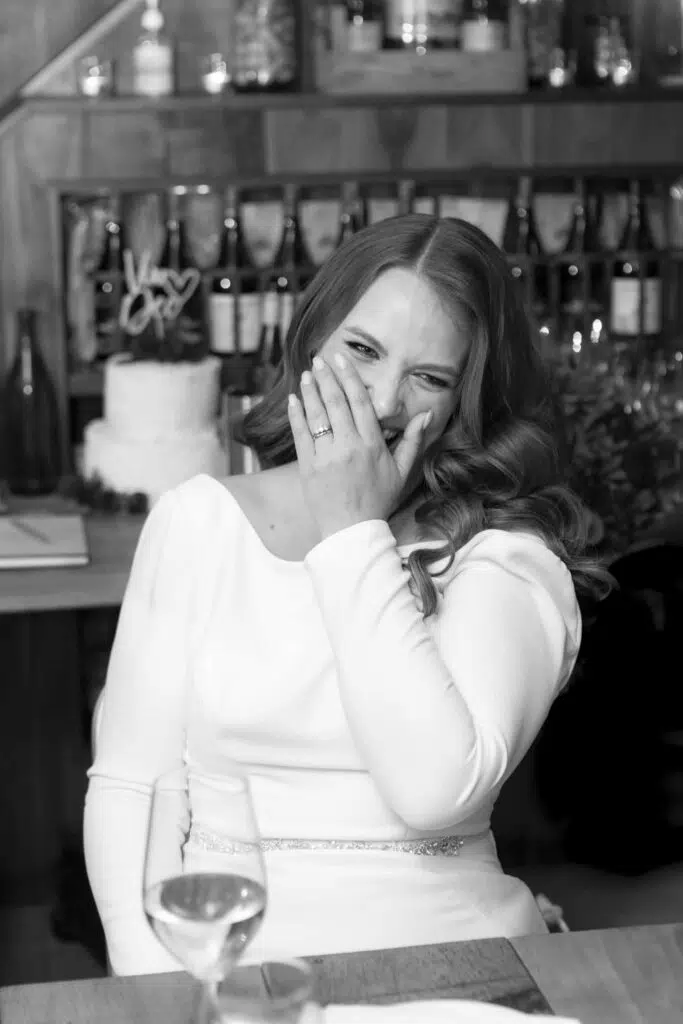 Black and white photograph of a bride covering her mouth as she laughs during her wedding reception in London
