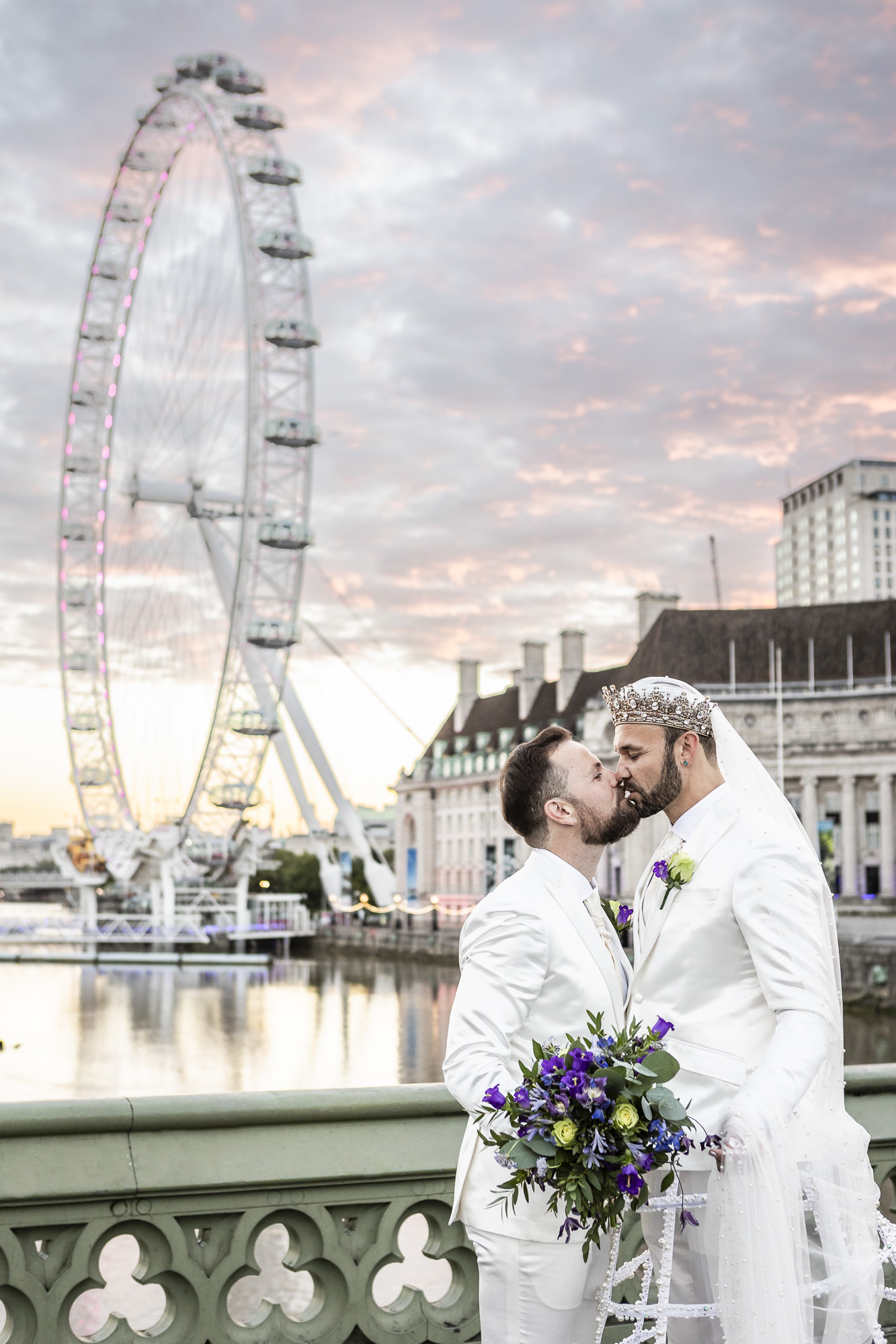 Two grooms kissing on Westminster Bridge at dawn, with the London Eye and soft pastel sky behind them during their Chelsea Old Town Hall LGBTQ+ micro wedding.